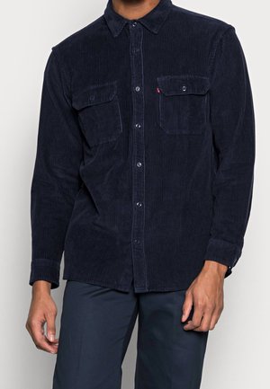 Mężczyzna w ciemnogranatowej sztruksowej koszuli na guziki z dwoma kieszeniami na piersi i długimi rękawami, w połączeniu z ciemnymi spodniami.