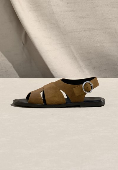 Καφέ σουέντ φλατ σανδάλι με ανοιχτά δάχτυλα, φαρδιές τιράντες, μαύρη σόλα και μεγάλο ασημί αγκράφα σε μπεζ υφασμάτινο φόντο.