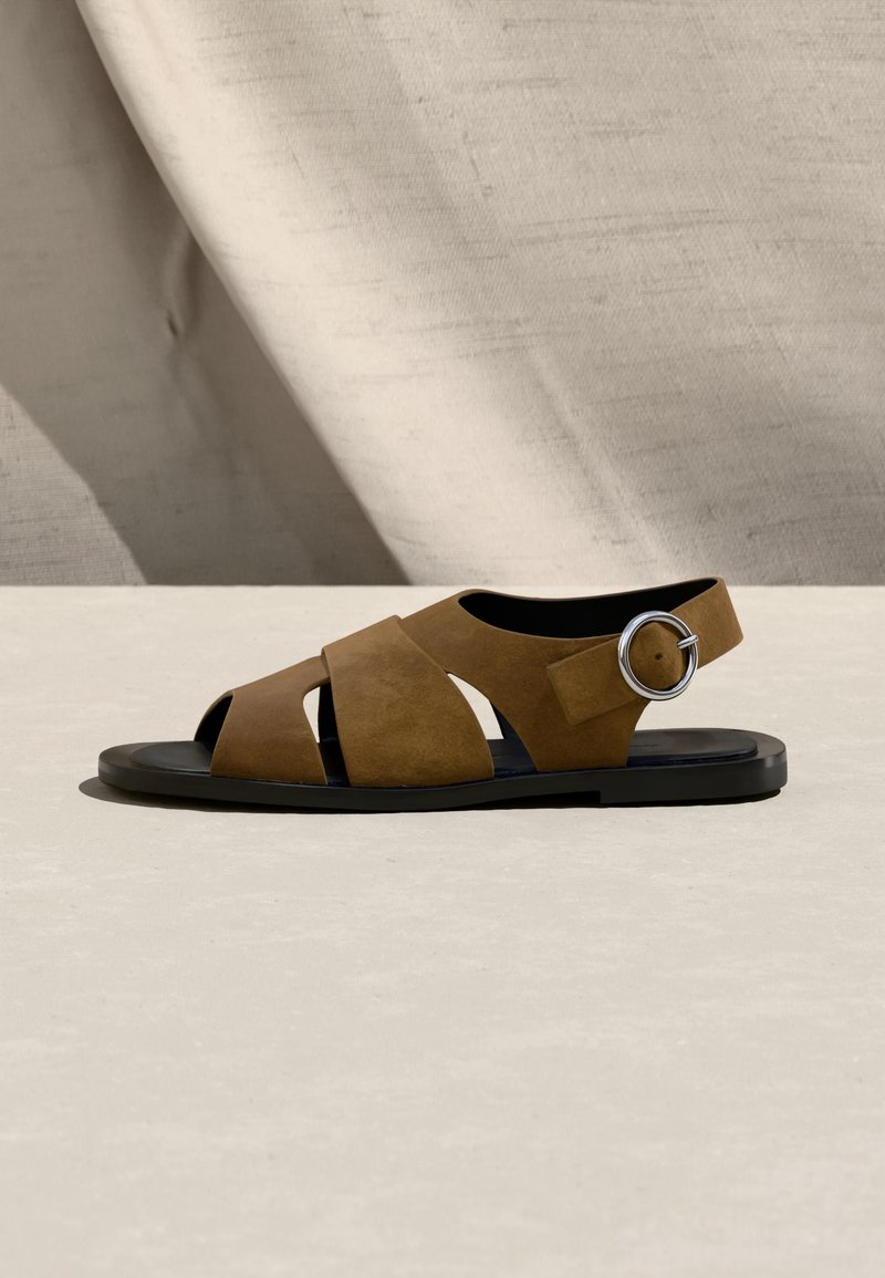 Sandalia plana de ante marrón con punta abierta, tiras anchas, suela negra y hebilla grande plateada sobre un fondo beige texturizado.