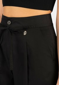 I pantaloni neri a vita alta presentano una chiusura con bottone, un nodo frontale e un accento in metallo con il logo. Il tessuto è liscio e presenta pieghe laterali.