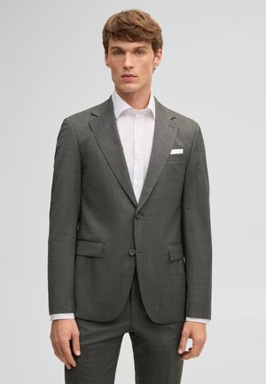 Giovane uomo con un abito grigio aderente, camicia bianca e fazzoletto bianco nel taschino, in piedi davanti a uno sfondo grigio uniforme.