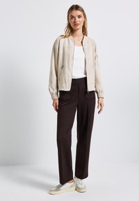 Beżowa kurtka bombowiec z zamkiem z przodu i elastycznymi mankietami, zestawiona z ciemnobrązowymi spodniami o szerokich nogawkach i jasnymi sneakersami. Gładka tekstura, minimalny design.