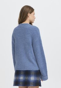 Maglione blu fluffy con una vestibilità rilassata, maniche lunghe e scollatura arrotondata; indossato sopra una gonna a quadri con tonalità blu e grigie.
