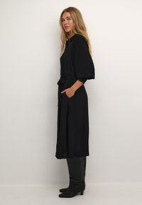 Zwarte lange jurk met een strik taille, korte pofmouwen, zijzakken, een gladde textuur en een losse pasvorm. Gecombineerd met zwarte knielange laarzen.