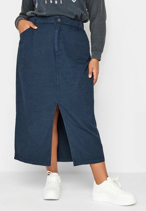 Person iført en navyblå midinederdel med frontslids, en mørk langærmet top og hvide sneakers, stående med den ene hånd i lommen.
