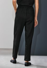 Pantaloni negri conici cu talie elastică, având o textură netedă și tivuri întoarse. Purtați cu pantofi negri slip-on.