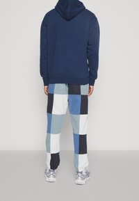 Sudadera azul marino con capucha y bolsillo tipo canguro combinada con pantalones deportivos de patchwork en cuadros azules, blancos y azul marino. Zapatillas grises completan el conjunto.
