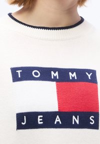 Gros plan d’un pull en maille blanc avec un logo rectangulaire bleu marine, blanc et rouge et le texte "TOMMY JEANS" sur la poitrine.