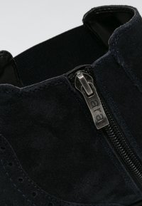Primer plano de una bota de tobillo de ante negro con tira de metal grabada "ara" y panel lateral elástico.