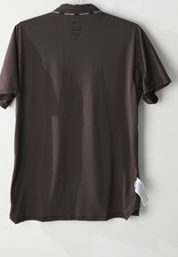 Hnedé tričko s krátkym rukávom Nike Dri-FIT s brandingom NikeCourt vo vnútri goliera, zavesené na bielej stene, ukazujúce zadnú stranu a etiketu s pokynmi na údržbu.