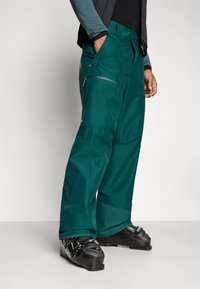 Tyrkysové nepremokavé lyžiarske nohavice s voľným strihom, zipsovými vreckami, spevnenými manžetami a odolným materiálom na zimné športy.