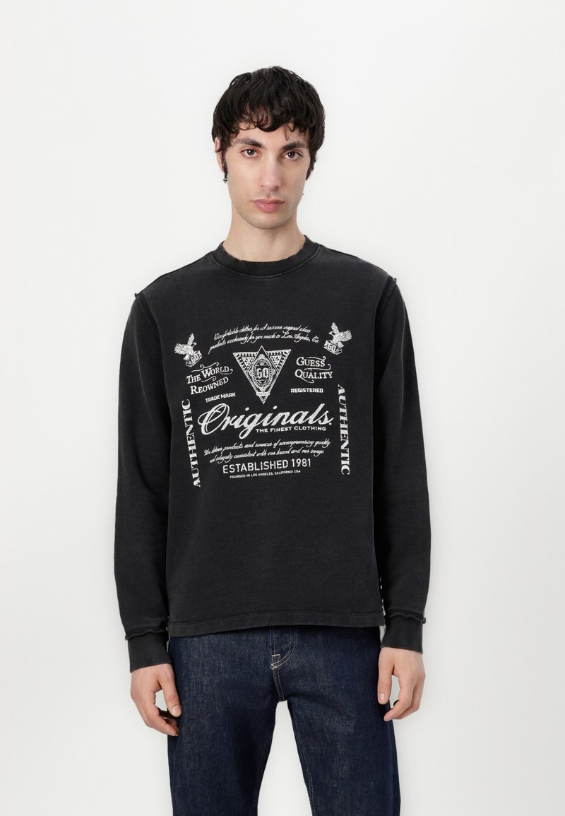 Felpa nera con grafiche stampate, caratterizzata da un collo e polsini a costine e una vestibilità leggermente oversize. Indossata con jeans di denim blu.