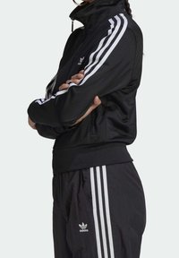 Svart sportjacka med dragkedja och hög krage, med vita tre-randiga detaljer på ärmarna och nederkanten, lättviktsmaterial, sportig design.