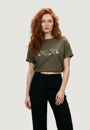 Mujer con cabello largo y rojo lleva una camiseta corta verde oliva con estampado floral y pantalones de talle alto negros contra un fondo liso.