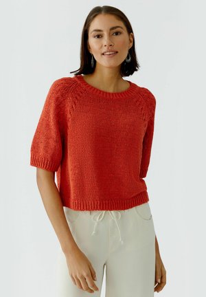 Vrouw draagt een rood gebreide korte mouwen trui met crèmewitte broek met trekkoord, staand tegen een effen lichte achtergrond.