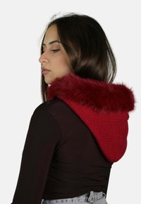 Felpa rossa lavorata a maglia con cappuccio in pelliccia sintetica, che presenta un design strutturato e un colore vivace, completata da una semplice maglietta nera e jeans.