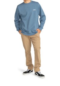 Sudadera de cuello redondo azul con logo blanco, pantalones beige y zapatillas negras. La sudadera tiene un corte holgado y puños y dobladillo acanalados.