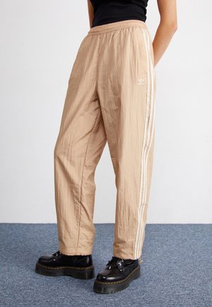 Person trägt beige Adidas-Trainingshose mit weißen Streifen und schwarzen Plateauschuhen und steht auf blauem Teppichboden.