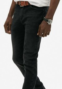 Vaqueros de denim negros con un corte ajustado, que cuentan con un cierre clásico de botón y cremallera. Combinados con un cinturón de cuero marrón y un reloj.