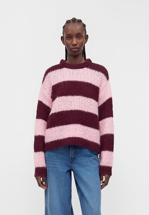 Femme aux cheveux tressés portant un pull en maille épaisse avec de larges rayures horizontales roses et bordeaux et un jean bleu, debout devant un fond uni.