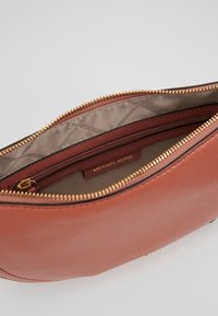 Brun lædertaske med en toplynlås. Indvendigt har den en beige foring og en lille lynlåslomme med teksten "MICHAEL KORS." Hardware i guldfarve.