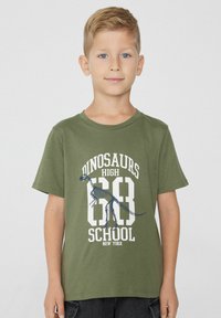Zelené bavlněné tričko s krátkými rukávy a kulatým výstřihem. Na přední straně je grafika dinosaurího kostry a bílý text „DINOSAURS HIGH SCHOOL 63 NEW YORK.“