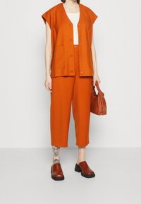 Oranžový dvoudílný outfit, který zahrnuje krátký rukáv, košilový top s kapsami a široké kalhoty s kratšími nohavicemi. Doplňky zahrnují hnědou kabelku a dřeváky.