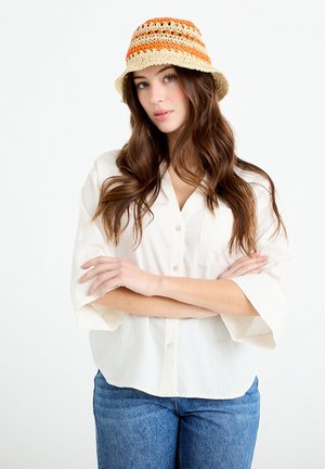 Frau mit langen braunen Haaren, die eine orange-beige gestreifte gehäkelte Mütze, ein weißes Hemd mit Knöpfen und blaue Jeans trägt, mit verschränkten Armen stehend.