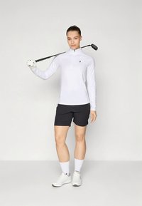 White long-sleeve golf top with a half-zip collar, paired with black shorts. The model holds a golf club over her shoulder, wearing white socks and shoes.