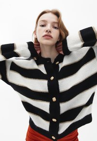 Svart och vit randig cardigan med guldiga detaljer, ribbad struktur och knappstängning. Har vida ärmar och en kortad design.