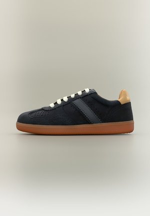Baskets en suède noir avec des lacets blancs, semelle en caoutchouc marron et languette de talon beige montrant une partie d’un logo, vue de côté sur fond uni.