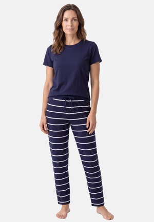 Vrouw staat blootsvoets, draagt een marineblauw T-shirt met korte mouwen en marineblauwe broek met witte horizontale strepen en een koord in de taille.
