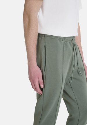 Pantalones de chándal verdes con cintura ajustable, bolsillos laterales y una textura suave. Llevados con una camiseta blanca, mostrando una vista lateral.