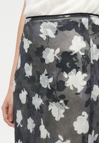 Τμήμα μέσης και φούστας με μαύρο, λευκό και γκρι λουλουδένιο μοτίβο, φορεμένο με λευκό τοπ, με το αριστερό χέρι να ακουμπά δίπλα στο σώμα.