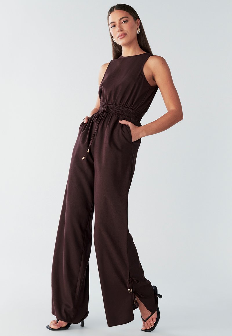 Rudonos spalvos be rankovių kombinezonas su elastine juosmens juosta, šoninėmis kišenėmis ir platėjančiomis kelnėmis; dekoruotas auksinėmis detalėmis ir tekstūruota medžiaga.