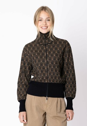 Giovane donna sorridente, indossa un maglione con zip e motivo, con polsini e orlo a costine neri e pantaloni beige su uno sfondo semplice.