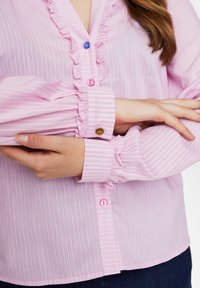 Blusa a righe rosa con colletto arricciato, dotata di bottoni rosa, blu e marroni e maniche arricciate, che mostra una texture di tessuto leggero.