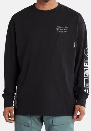 Man draagt een zwart Timberland shirt met lange mouwen, met witte tekst en symbolen op het borstzakje en de rechtermouw, gecombineerd met teal cargobroek.