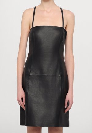 Vrouw die een mouwloze zwarte leren jurk met dunne schouderbandjes draagt, staand tegen een effen lichte achtergrond.