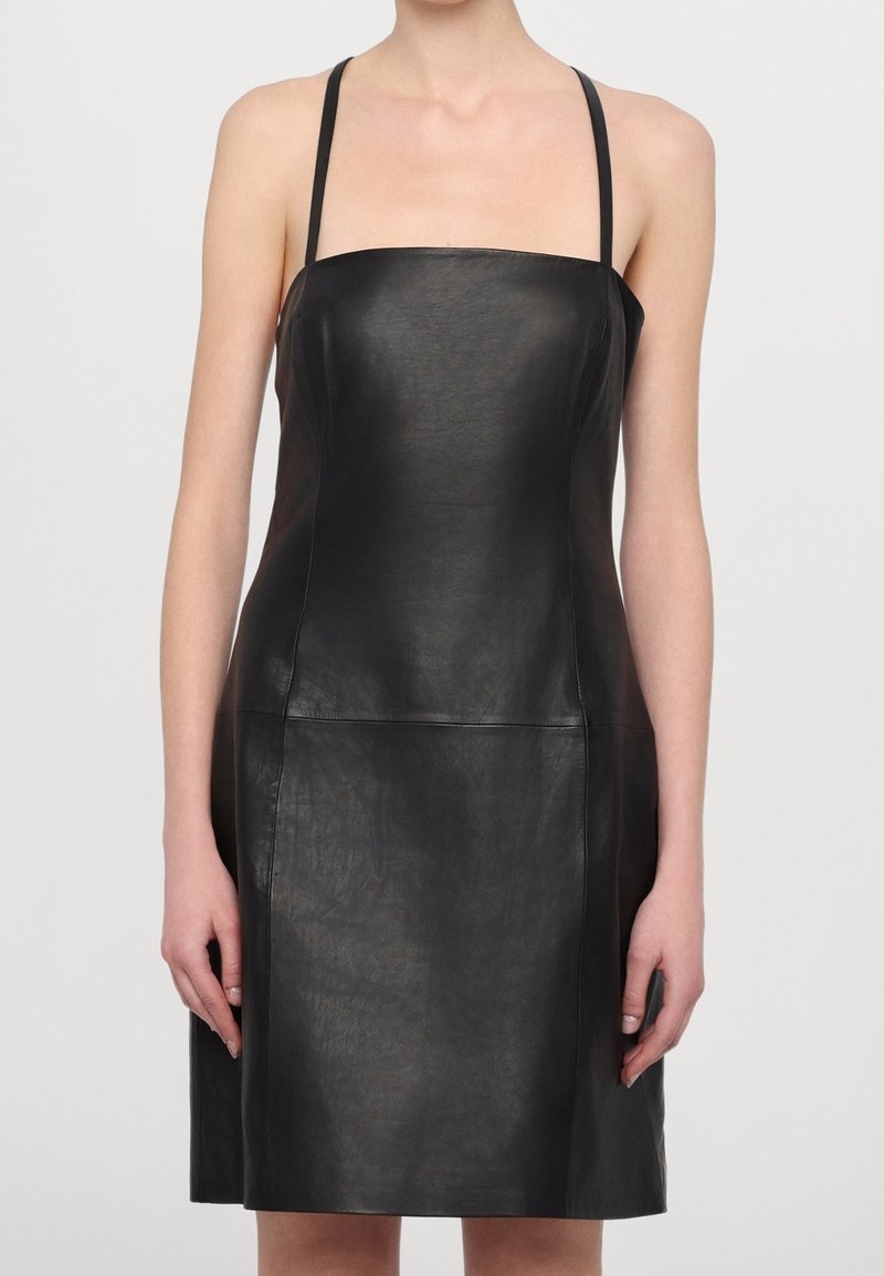 Femme portant une robe en cuir noire sans manches avec de fines bretelles, debout devant un fond clair uni.