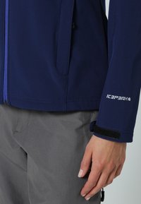 Chaqueta de softshell de color marino con cierre frontal de cremallera, bolsillos laterales y puños de velcro. Confeccionada en una tela suave con una textura ligera.