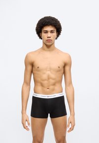 Μαύρα μποξέρ με λευκή ελαστική μέση που φέρει το λογότυπο "TOMMY HILFIGER". Το μοντέλο έχει καλογυμνασμένο σώμα και κοντά, σγουρά μαλλιά.