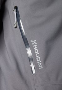 Šedá softshellová bunda so zipsovým vreckom, reflexným lemovaním a diskrétnym logom. Materiál sa zdá byť ľahký a hladký.
