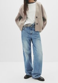 Femme portant un cardigan beige sur une chemise blanche, un jean ample bleu clair et des mocassins noirs, se tenant devant un fond blanc uni.