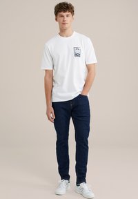 Balta, trumpa rankovė t-shirt su mažu mėlynu grafiniu elementu kairėje krūtinės pusėje, derinama su tamsiai mėlynomis džinsomis ir baltais sportbačiais. Neutrali fono spalva.