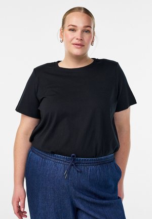 Nuori nainen, jolla on vaaleat hiukset sidottuna taakse, pukeutuneena mustaan lyhythihaiseen paitaan ja sinisiin naruhousuihin, seisoo valkoista taustaa vasten.