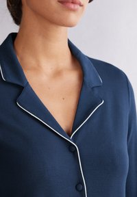 Mörkblå pyjamasöverdel med en slät textur, med en notchkrage och vit kantad detalj längs kanterna, knäppt med två knappar.
