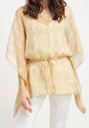 Kvinde iført en løs, beige bluse med slangeprint, brede ærmer og snøre i taljen, kombineret med hvide bukser.