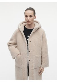 Cappotto in pile beige con cappuccio, dotato di due tasche frontali, chiusura a bottoni e una finitura morbida e testurizzata. Indossato sopra un maglione nero.
