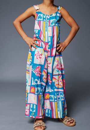 Enfant portant une combinaison sans manches colorée avec des motifs de dessins animés et des sandales roses, debout les mains sur les hanches devant un fond gris.